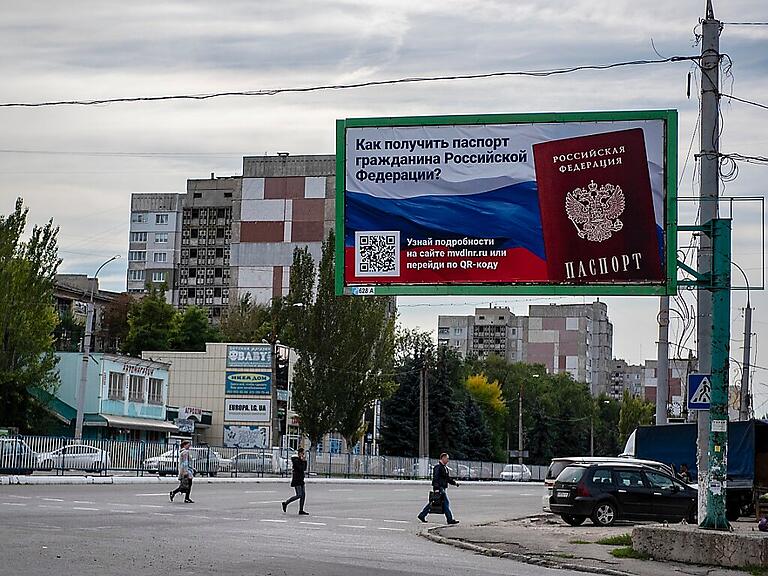 Menschen überqueren eine Straße mit einem Plakat mit der Aufschrift "Wie man den Pass eines russischen Bürgers bekommt" vor dem Scheinreferendum in der von den Russen kontrollierten Volksrepublik Luhansk. Foto: Uncredited/AP/dpa