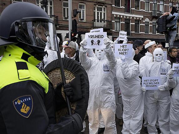 Demonstranten halten Plakate hoch, während ein Bereitschaftspolizist im Vordergrund steht. In Amsterdam hat die Polizei eine verbotene Demonstration gegen die Corona-Politik der Regierung aufgelöst..Am Sonntag hatten nach Angaben der Nachrichtenagentur ANP rund 2000 Menschen demonstriert. Foto: Peter Dejong/AP/dpa